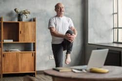 A middle aged white male is in a modern office doing knee stretches behind his desk.