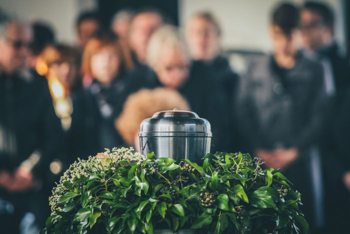 A metal urn in the foreground with a group of mourners behind it.