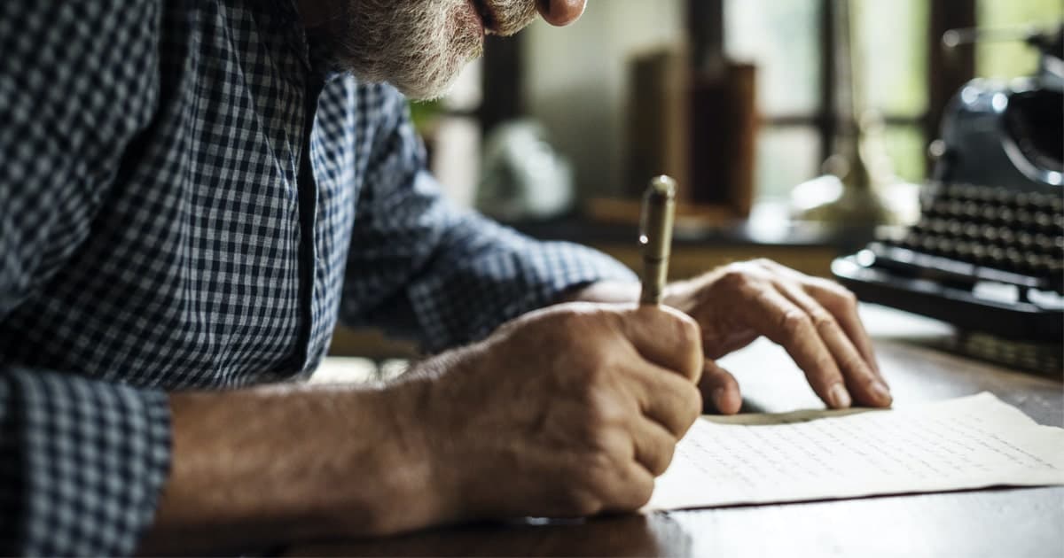 Man bent over a desk handwriting a letter on white paper.