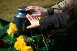 Two mourners' hands are touching a black urn amid yellow flowers.