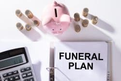 A collection of items including a piggy bank, rolls of coins, a calculator, and a binder opened to a page that reads "Funeral Plan."
