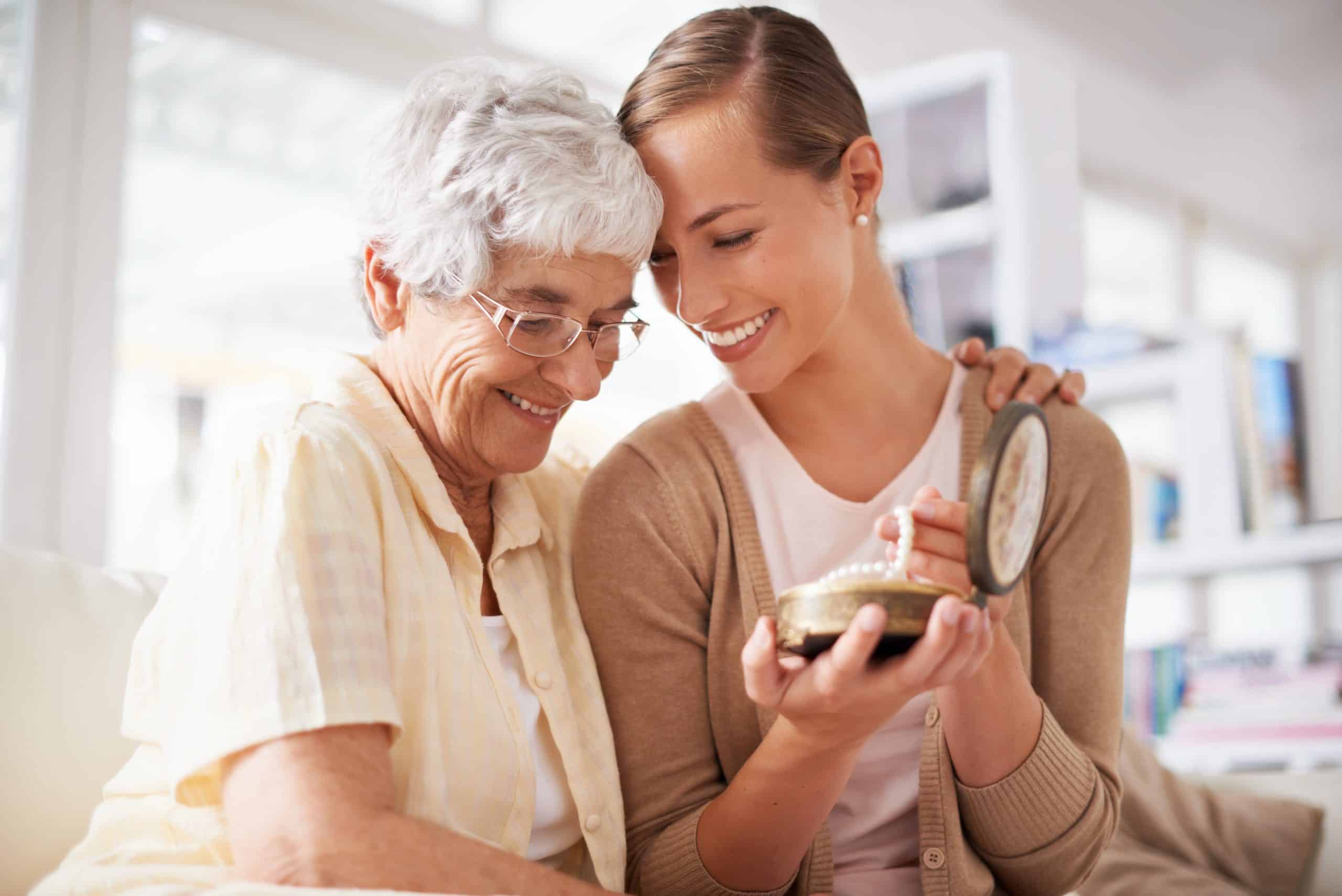 An older woman embraces a younger adult woman who is holding a string of pearls and a jewelry box.