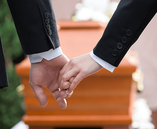 Two mourners in dark suits holding hands in front of a wooden casket at a funeral service