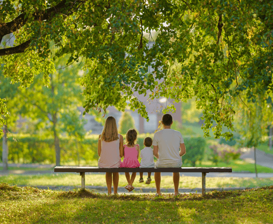 Parents and two young children sitting together on a park bench under a shade tree