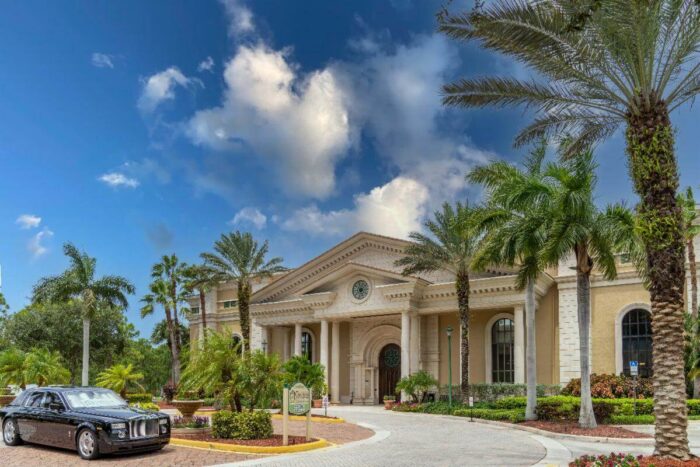 Entrance of The Gardens of Boca Raton, a columned building framed by palm trees, with a black sedan in the drive