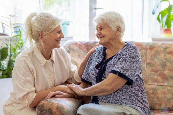 Adult daughter holding her elderly mother’s hand as they smile and talk on a living room sofa